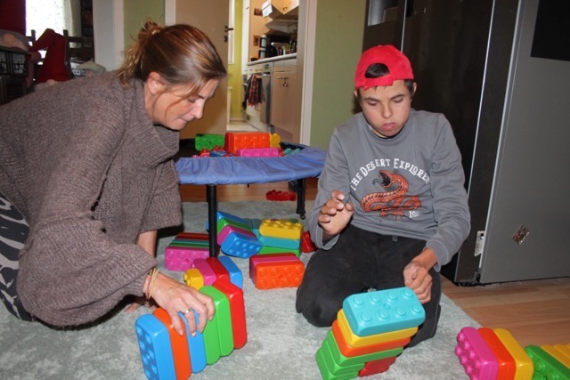 Eine Frau und ein Junge bauen gemeinsam mit bunten Playblocks auf einem Teppich. Vor ihnen stehen mehrere kleine Bauwerke.