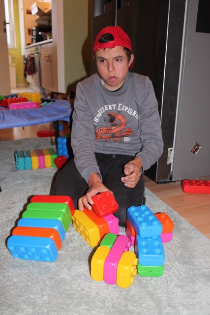 Ein junger Mann kniet auf dem Teppich und ordnet rote, blaue, grüne, gelbe und orangefarbene Playblocks zu verschiedenen Formen an.