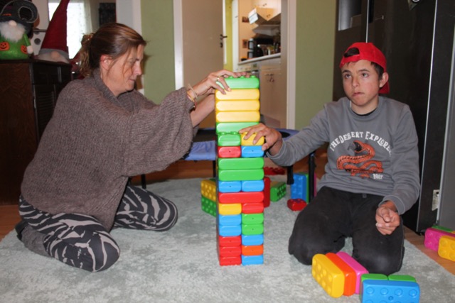 Eine Frau und ein junger Mann bauen gemeinsam einen hohen Turm aus bunten Playblocks und stabilisieren ihn mit den Händen.