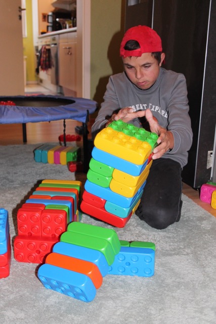 Ein junger Mann kniet vor mehreren Bauwerken und hält einen schräg gestapelten Turm aus bunten Playblocks fest.