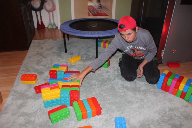 Ein junger Mann kniet auf einem Teppich und greift nach einem Bauwerk aus bunten Playblocks. Weitere Bausteine liegen im Raum verteilt.