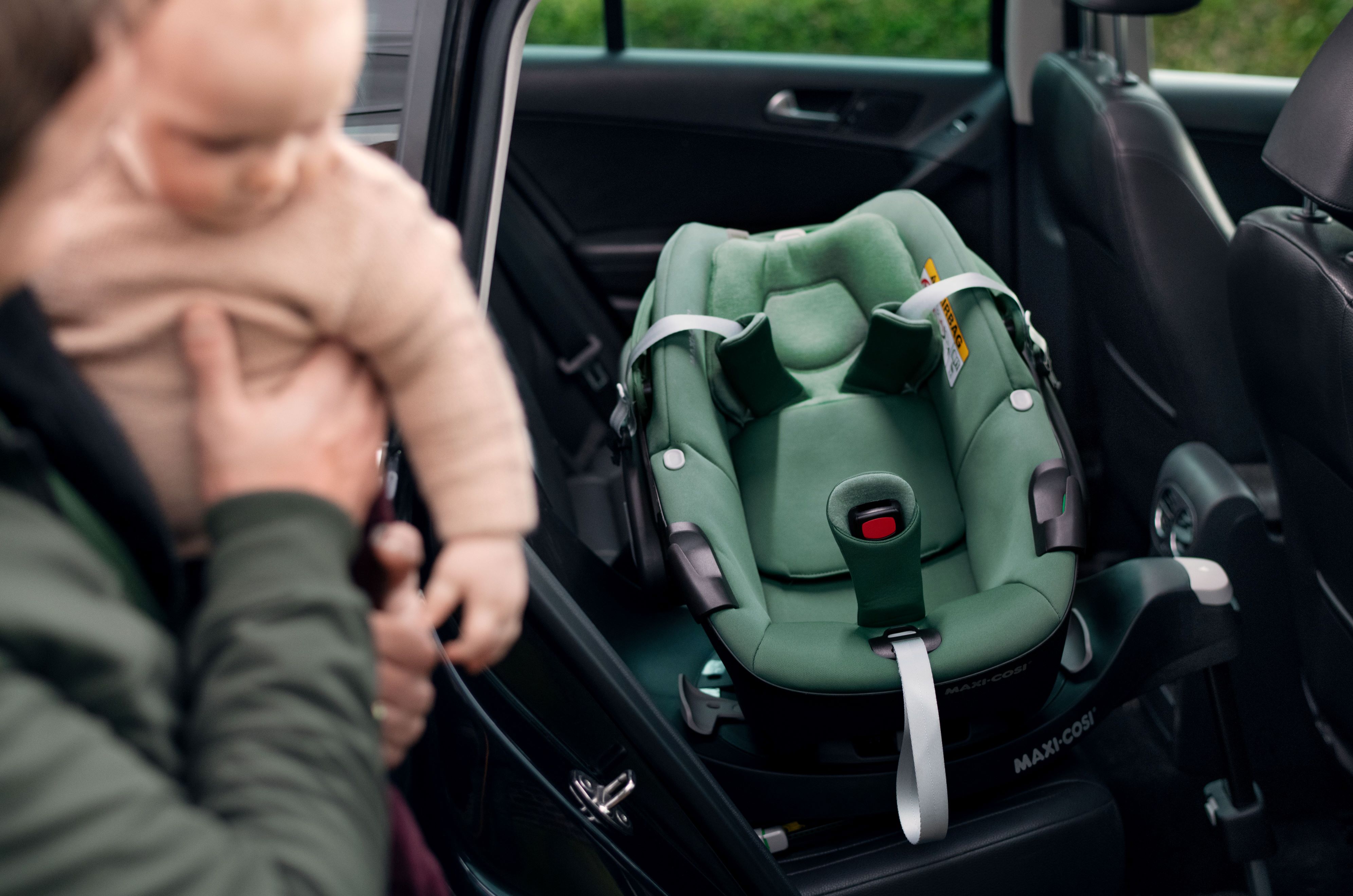 image Ein grüner Maxi-Cosi Kindersitz ist im Auto installiert, während ein Vater im Vordergrund sein Baby aus dem Fahrzeug hebt.