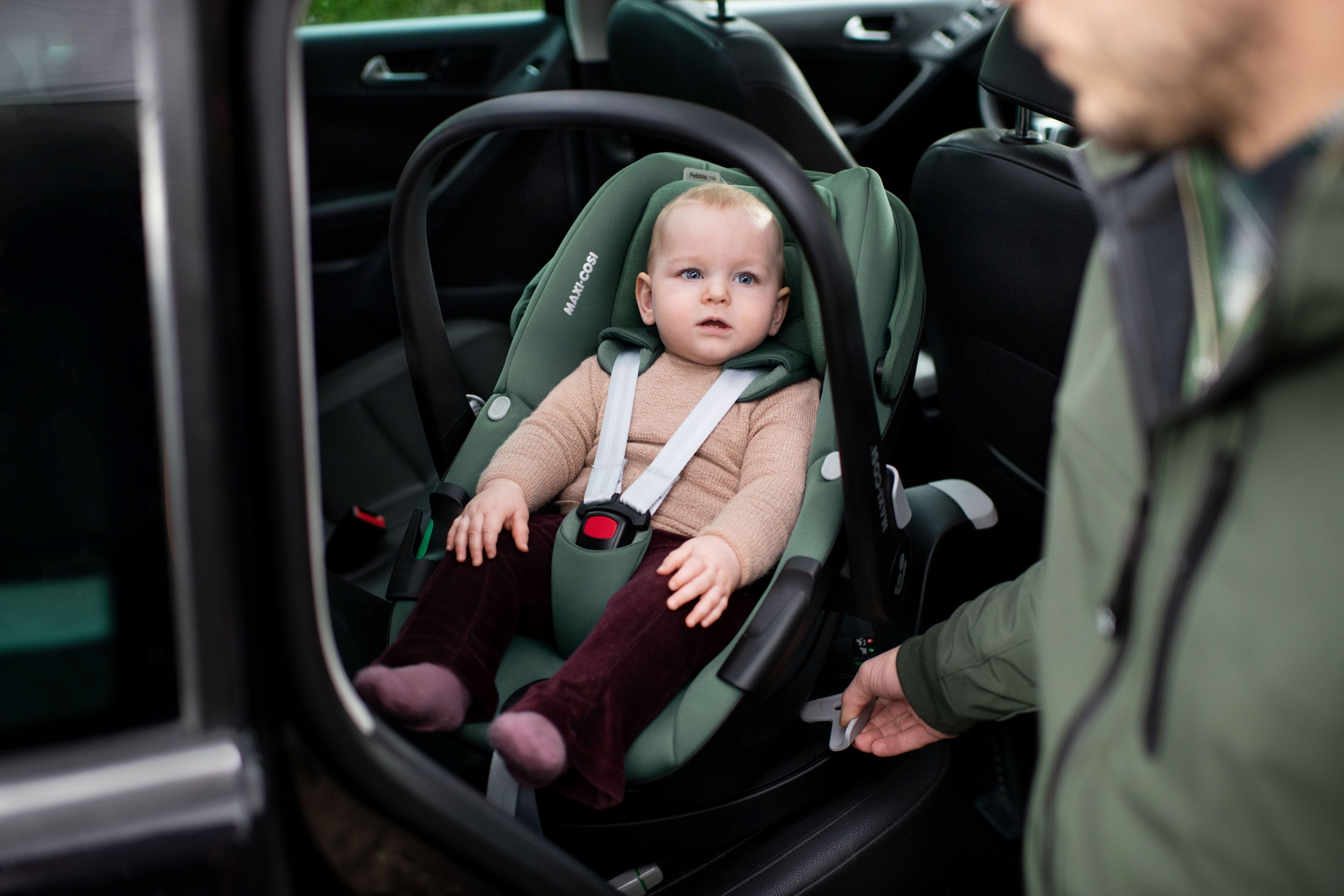 image Ein Baby sitzt in einer grünen Maxi-Cosi Babyschale im Auto, angeschnallt und aufmerksam blickend. Ein Erwachsener in grüner Jacke greift nach der Basisstation zum Lösen oder Befestigen der Babyschale.