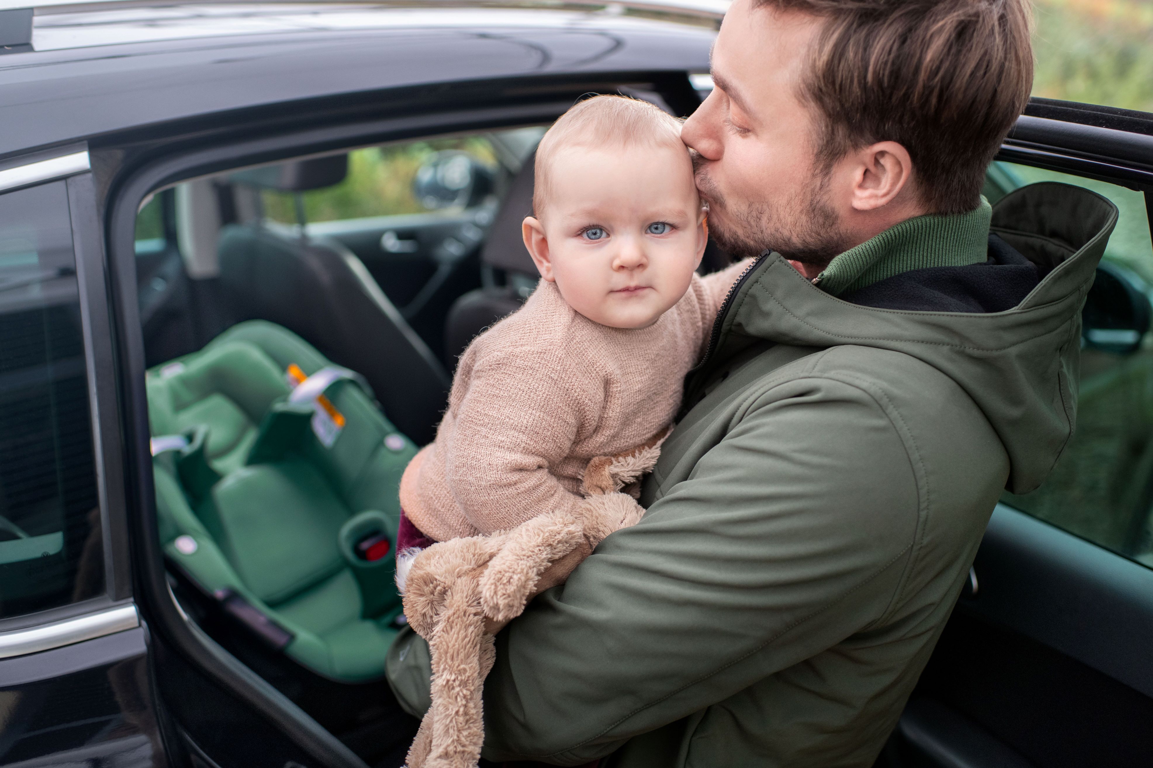image Vater hält Baby liebevoll im Arm und küsst es auf den Kopf, im Hintergrund ist eine Maxi-Cosi Babyschale im Auto zu sehen.