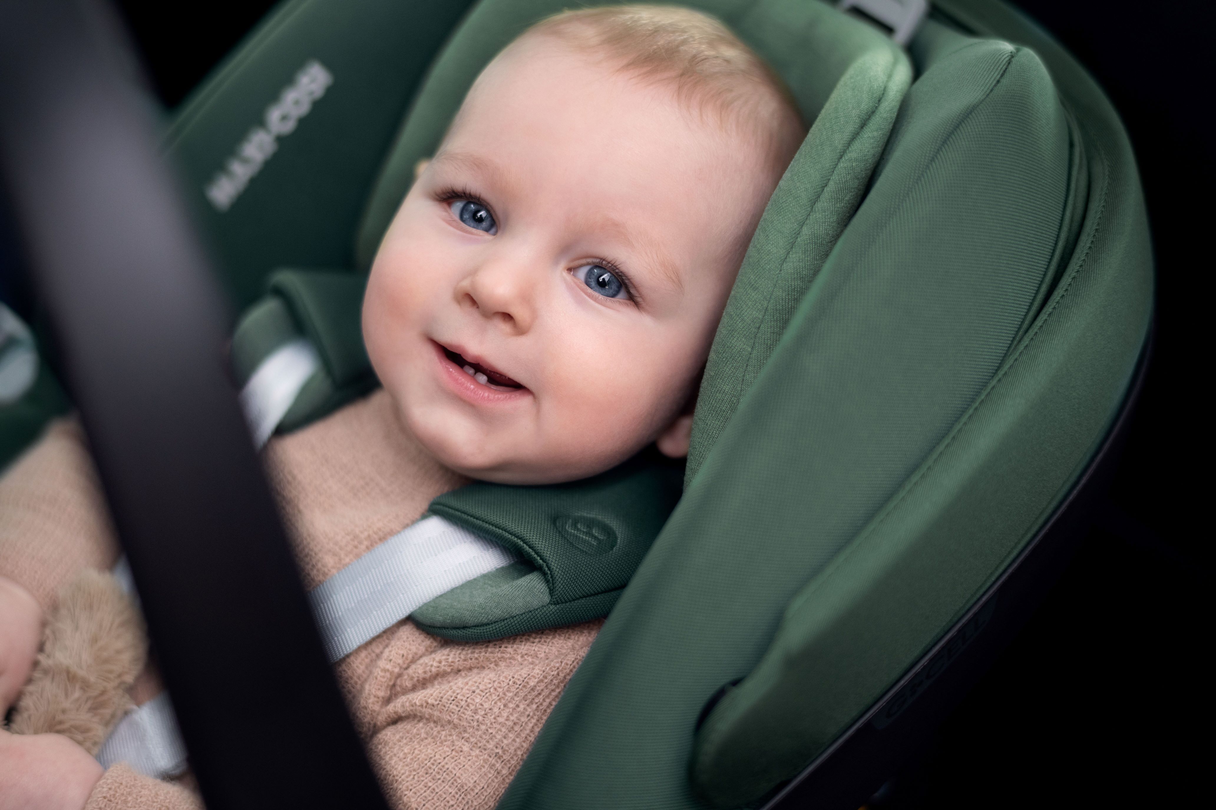 image Ein fröhliches Baby sitzt sicher angeschnallt in einem grünen Maxi-Cosi Kindersitz und lächelt mit großen blauen Augen in die Kamera.