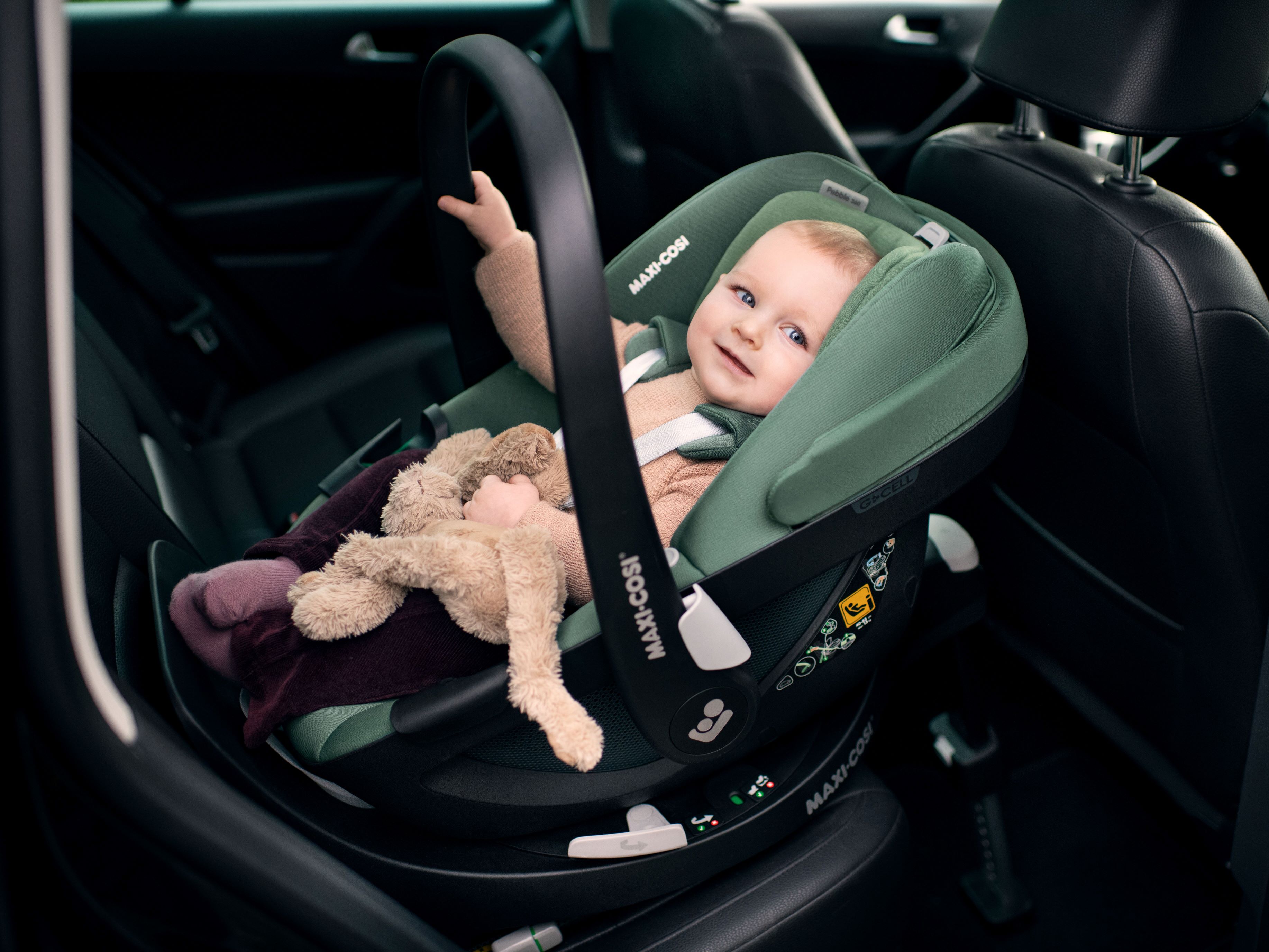 image Ein lächelndes Baby sitzt entspannt in einem grünen Maxi-Cosi Kindersitz auf der Rückbank eines Autos und hält ein Kuscheltier in der Hand.