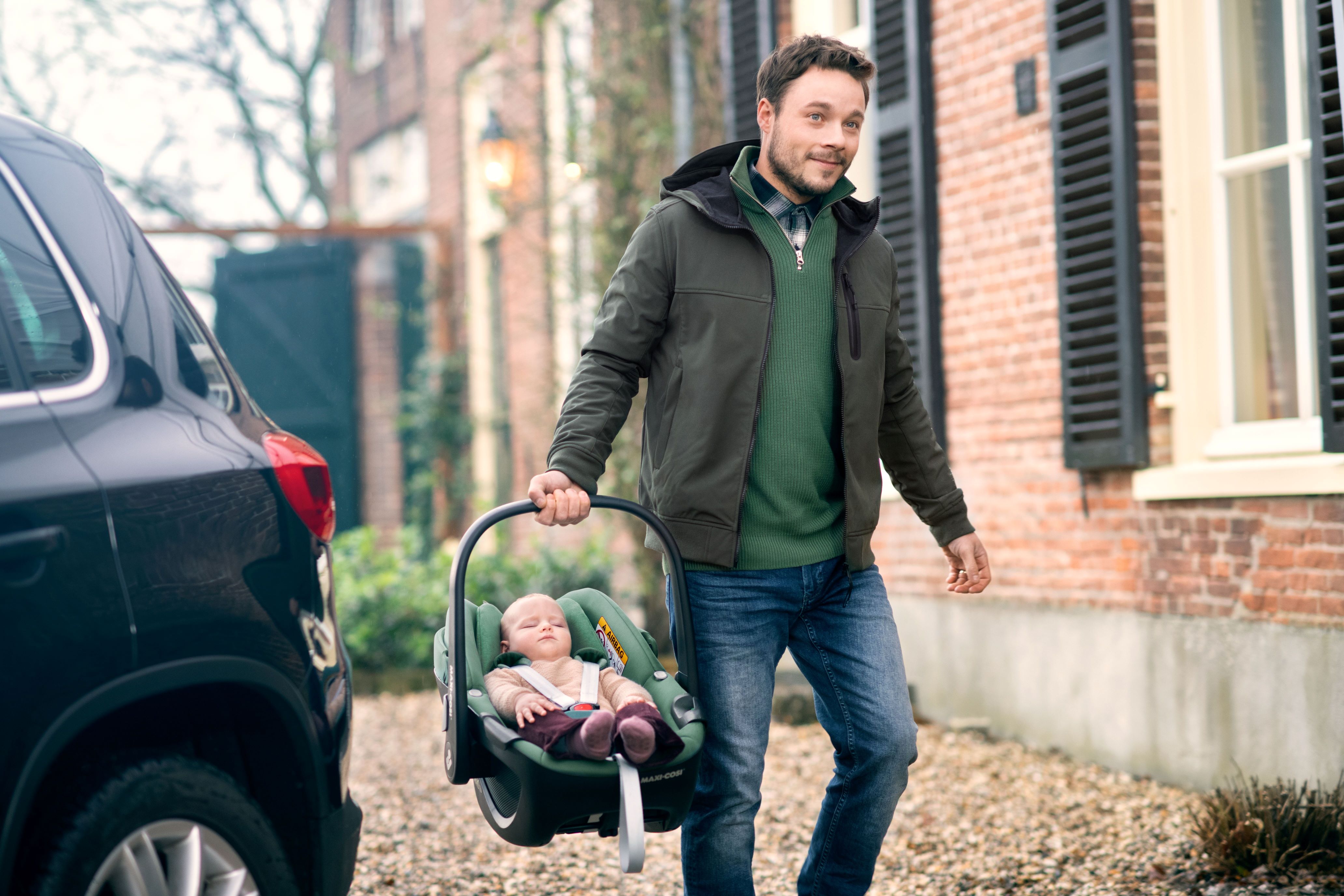 image Vater trägt Maxi-Cosi Babyschale mit entspanntem Baby darin über den Hof vor einem Wohnhaus.