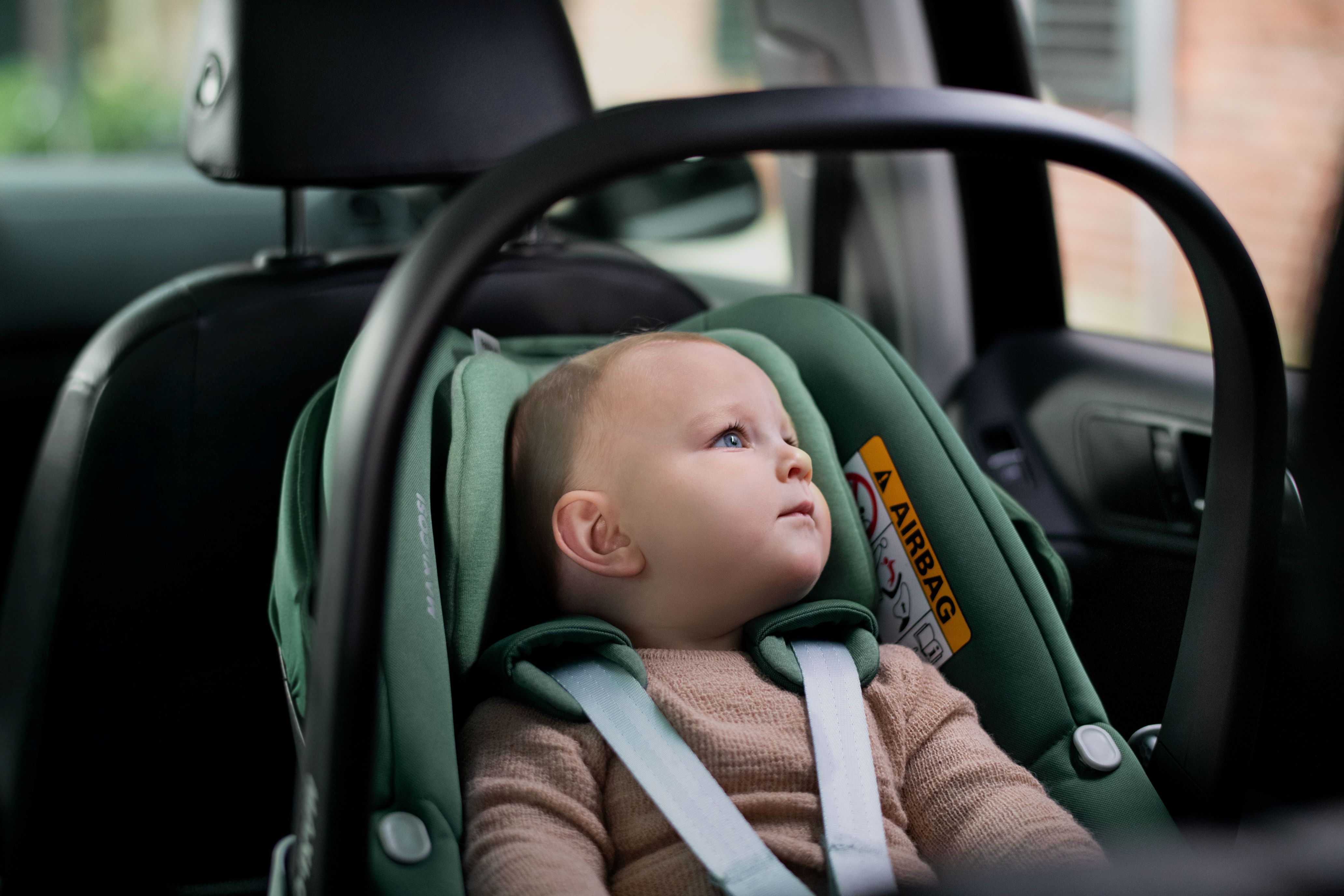 image Baby sitzt sicher angeschnallt in einer grünen Maxi-Cosi Babyschale im Auto und blickt neugierig nach oben.